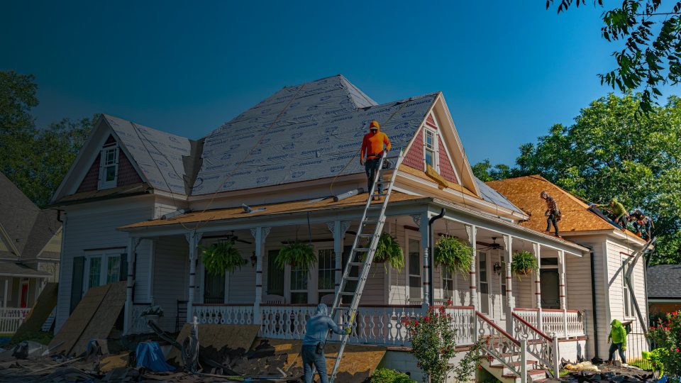 A crew of roofers working on a two-story home, installing synthetic underlayment and new plywood decking on a steep roof pitch using ladders and safety gear.