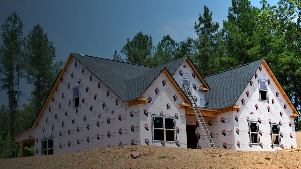 A new residential home under construction surrounded by trees, wrapped in protective house wrap with dark asphalt shingles installed across multiple roof gables.