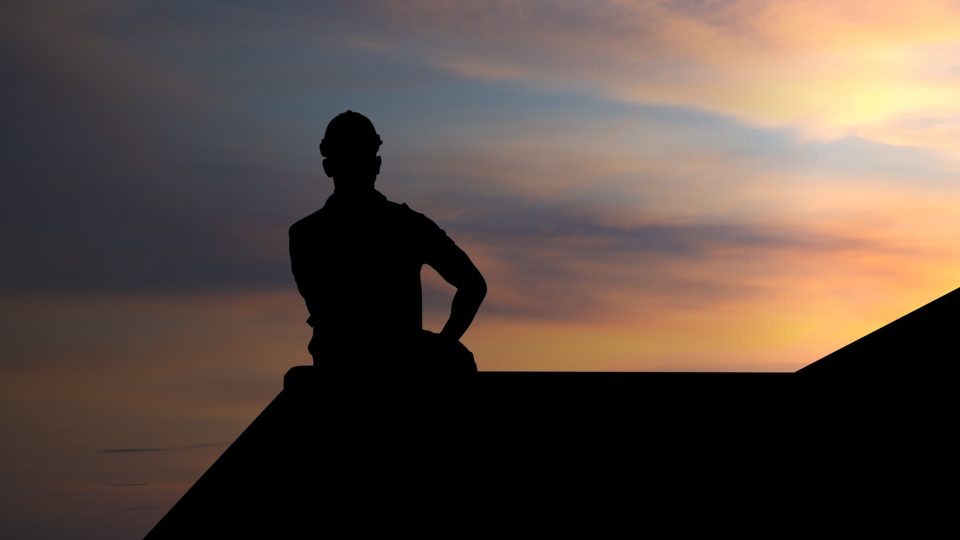 Dark silhouette of a worker wearing a hard hat perched on top of a roof peak, framed by a soft orange and purple sunset.